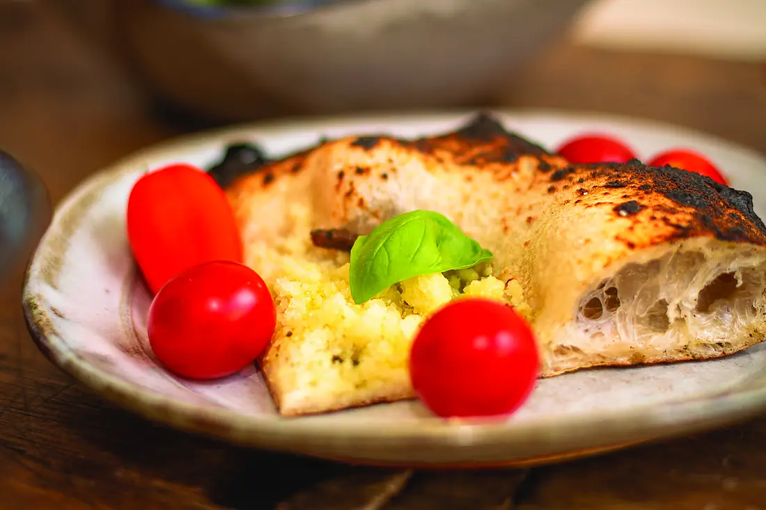 Fatia de pizza napolitana cortada ao meio, mostrando os alvéolos abertos da borda, ao lado de tomates.