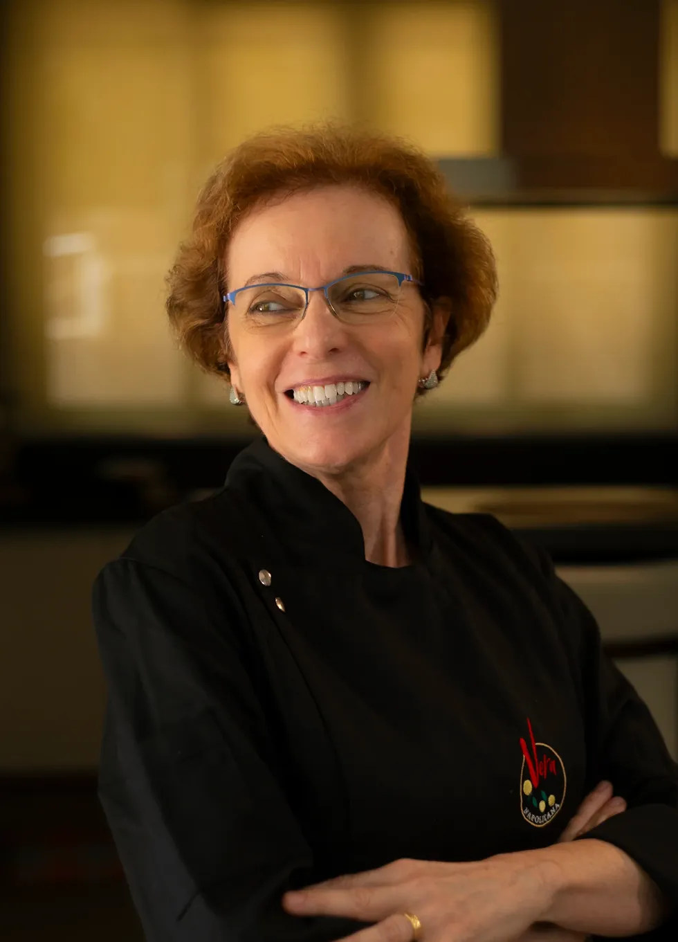 Vera Lucia Cegal, de dólmã preto e óculos, sorrindo de braços cruzados na cozinha.